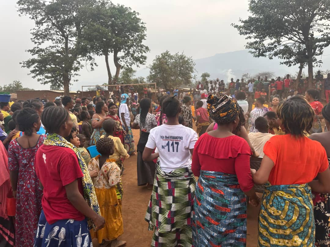 Rassemblement de villageois, surtout des femmes en pagnes colorés, sur une place de terre battue d'un village des hauts plateaux de Fizi, montagnes en arrière-plan ; au premier plan, une femme porte un t-shirt à l'effigie du député Obedi Nyamangyoku.