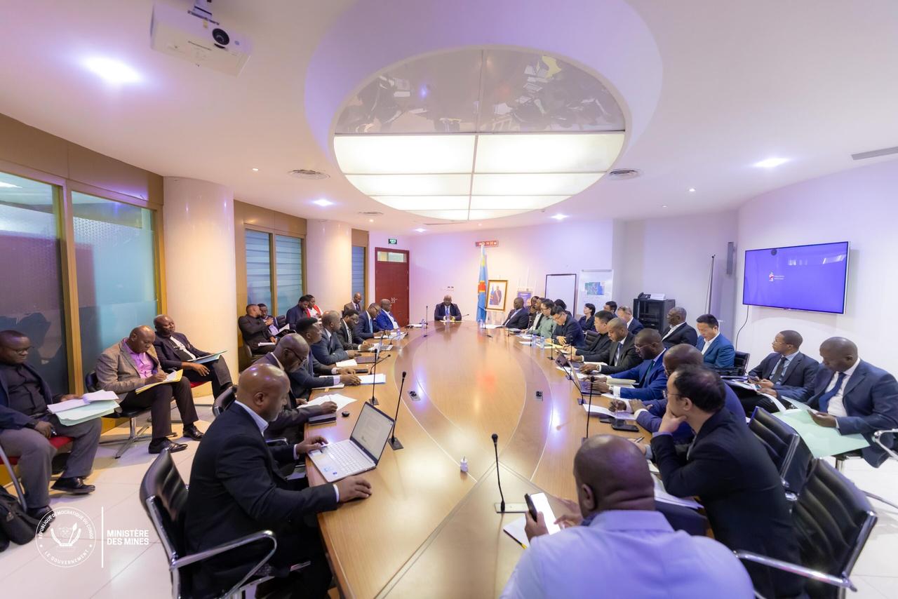 Vue d'ensemble d'une salle de réunion du ministère des Mines à Kinshasa, où des dizaines de représentants de sociétés minières sont assis autour d'une grande table ovale, devant le drapeau congolais et le portrait du chef de l'État.