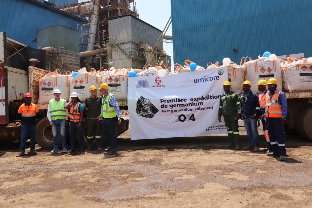 Des agents en tenue de travail posent devant une bannière STL, Gécamines et Umicore marquée « Première expédition de germanium », devant les big-bags de concentré chargés sur un camion et l'usine du Terril de Lubumbashi.