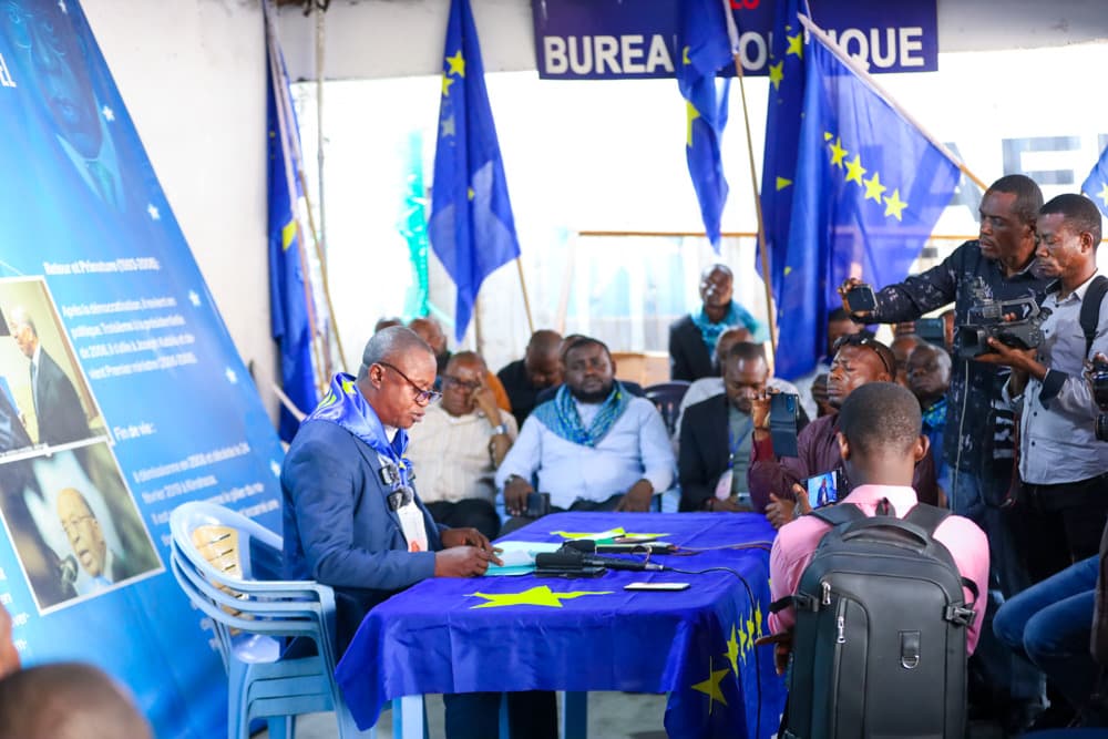 Un responsable du PALU lit une déclaration derrière une table drapée du drapeau bleu à étoiles jaunes du parti, entouré de drapeaux du PALU, de militants assis et de journalistes, sous une banderole « Bureau politique », au siège du parti à Kinshasa.
