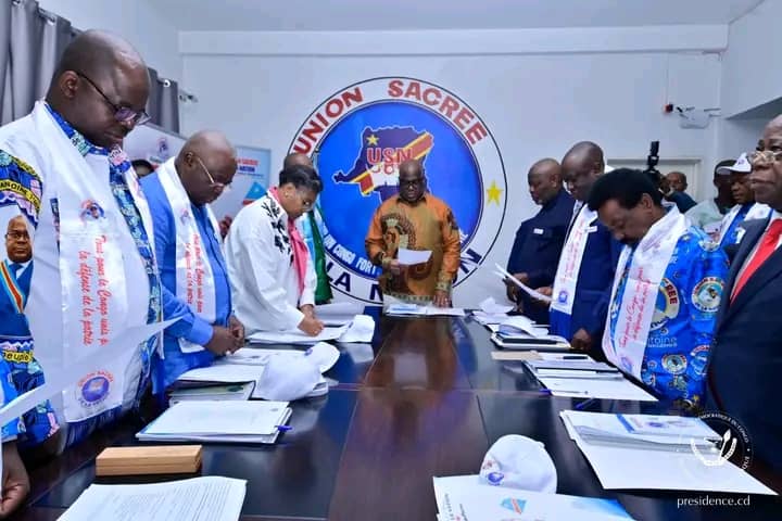 Des cadres de l'Union sacrée de la nation, écharpes aux couleurs de la plateforme, debout autour d'une table de réunion devant le logo de l'USN, à Kinshasa
