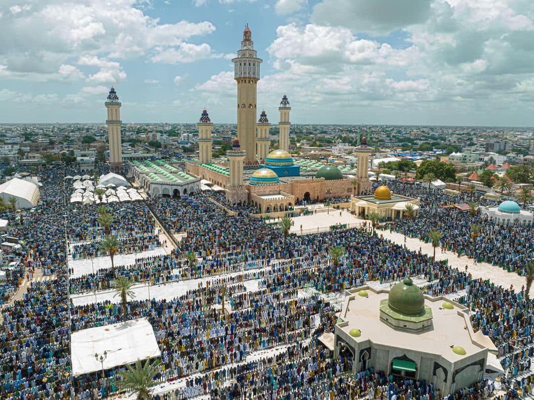 Vue aérienne de la grande mosquée de Touba, avec son grand minaret Lamp Fall et ses coupoles vertes et dorées, entourée d'une foule immense de pèlerins mourides rassemblés sur l'esplanade lors du Grand Magal.