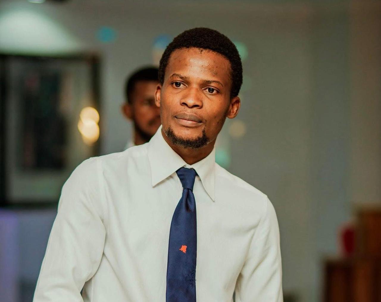 Portrait de Nathanaël Onokomba, chemise blanche et cravate bleu marine ornée d'une épinglette en forme de carte de la République démocratique du Congo