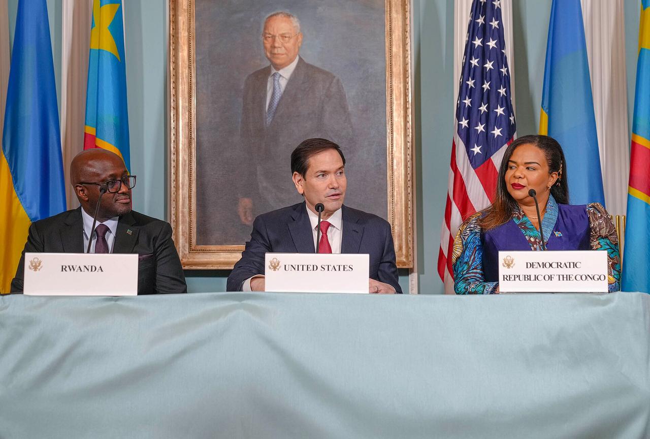 Trois responsables assis derrière une table recouverte d'une nappe bleue, devant des drapeaux du Rwanda, des États-Unis et de la République démocratique du Congo. Des plaques nominatives posées devant eux indiquent « Rwanda », « United States » et « Democratic Republic of the Congo ». À gauche, un homme en costume sombre portant des lunettes ; au centre, un homme en costume à cravate rouge qui parle dans un micro ; à droite, une femme en veste bleue et tenue en pagne coloré. Un portrait peint est accroché au mur derrière eux.