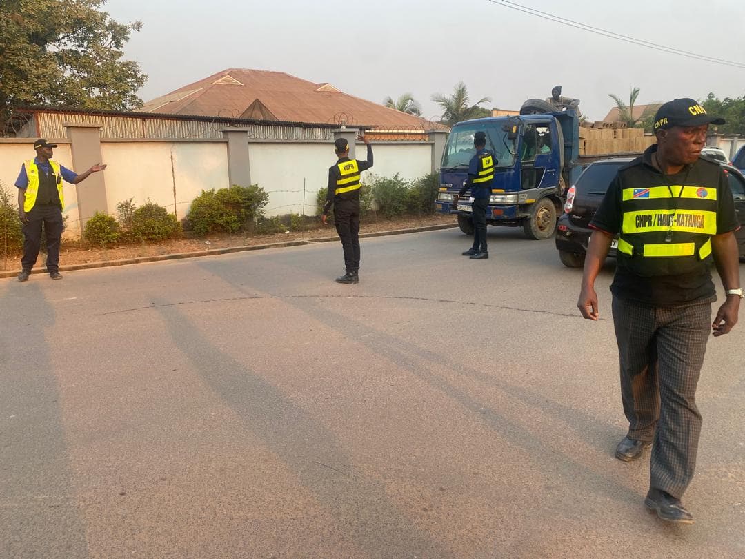 Des agents de la CNPR en gilets fluo régulent la circulation et un camion-benne sur une artère de Kamina, devant une villa au mur de clôture.