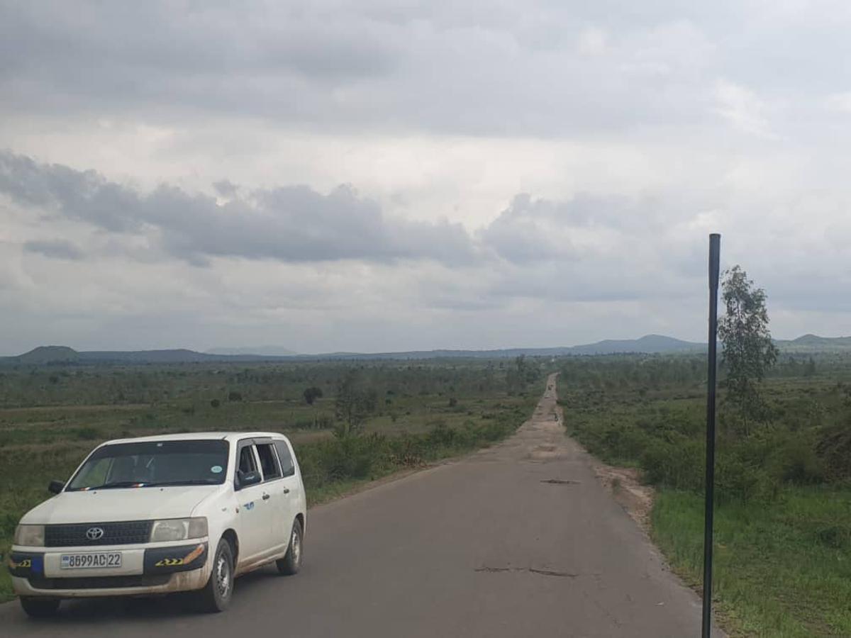Route bitumée traversant la plaine de la Ruzizi, dans le territoire d'Uvira, avec une camionnette blanche à gauche, des collines à l'horizon et un ciel nuageux.
