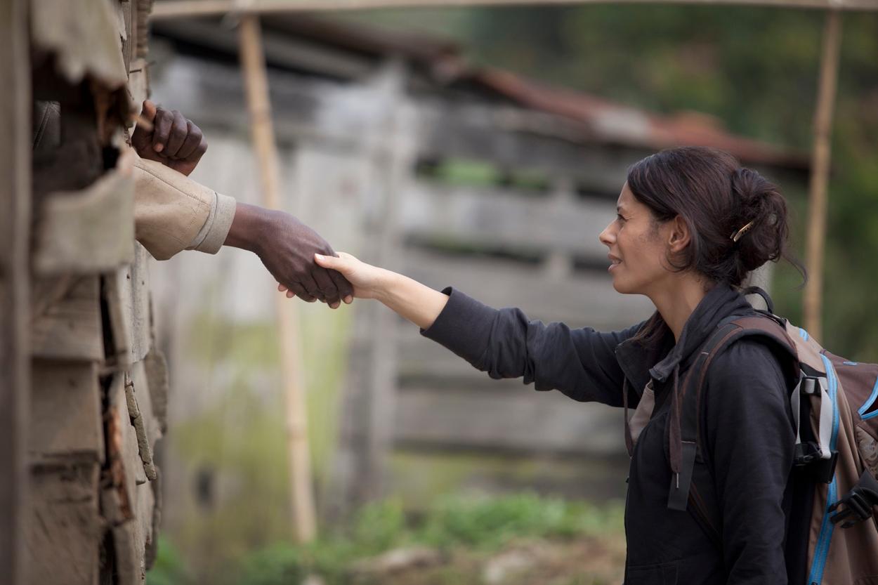 Une agente des droits de l'homme de la MONUSCO, sac à dos sur l'épaule, tend le bras et serre la main d'un détenu dont on ne voit que le bras et le poing sortant d'une ouverture dans le mur en planches de bois d'un lieu de détention.