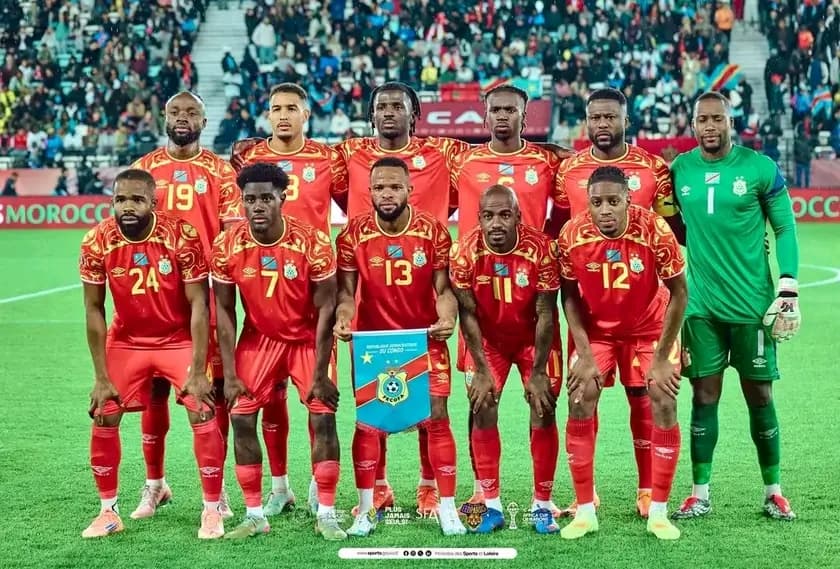Le onze de départ de la sélection congolaise de football, en maillot rouge, posant sur la pelouse d'un stade avant le coup d'envoi, le gardien en vert à droite.