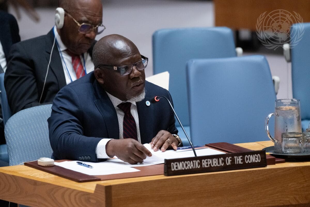 Un homme en costume sombre et lunettes, assis au Conseil de sécurité de l'ONU derrière une plaque « Democratic Republic of the Congo », s'exprime en tenant des documents, sous le logo des Nations unies.