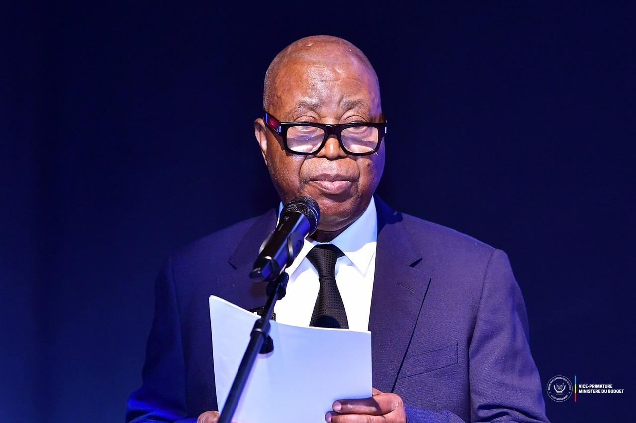 Adolphe Muzito, vice-Premier ministre au Budget, lit son discours au micro, une feuille à la main, sur une scène sombre marquée du logo de la Vice-Primature / Ministère du Budget.