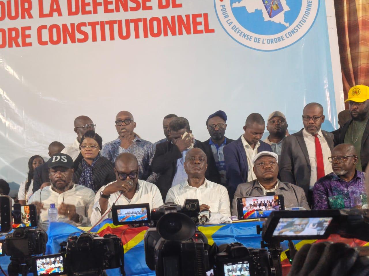Les responsables de la Coalition Article 64 assis en conférence de presse derrière une table drapée du drapeau de la RDC, l'un d'eux lisant une déclaration au micro, sous une grande banderole blanche « pour la défense de l'ordre constitutionnel » et le logo C64 ; des caméras et téléphones filment la scène au premier plan.