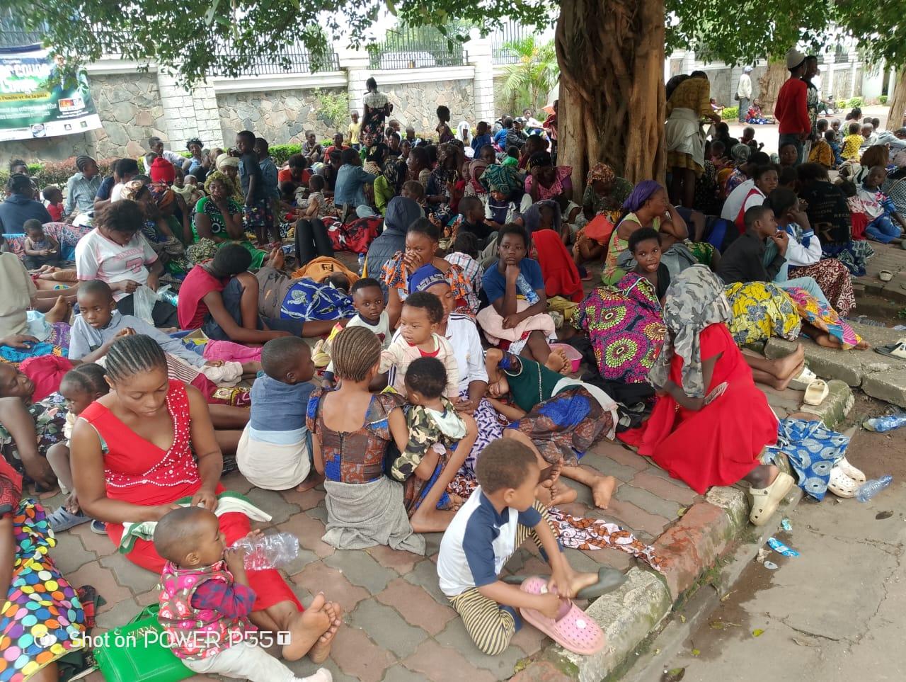 Des familles déplacées de l'Est, femmes et enfants, assises à même le sol devant le mur du gouvernorat du Haut-Katanga à Lubumbashi, une banderole de leur collectif visible en arrière-plan.