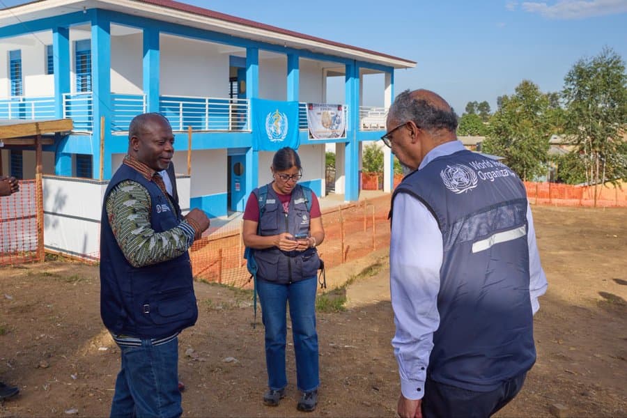 Deux responsables de la riposte et une agente de l'OMS en gilet bleu échangent dans la cour devant le bâtiment bleu et blanc du centre de traitement Ebola de Bunia, en Ituri, où flotte une banderole de l'OMS.