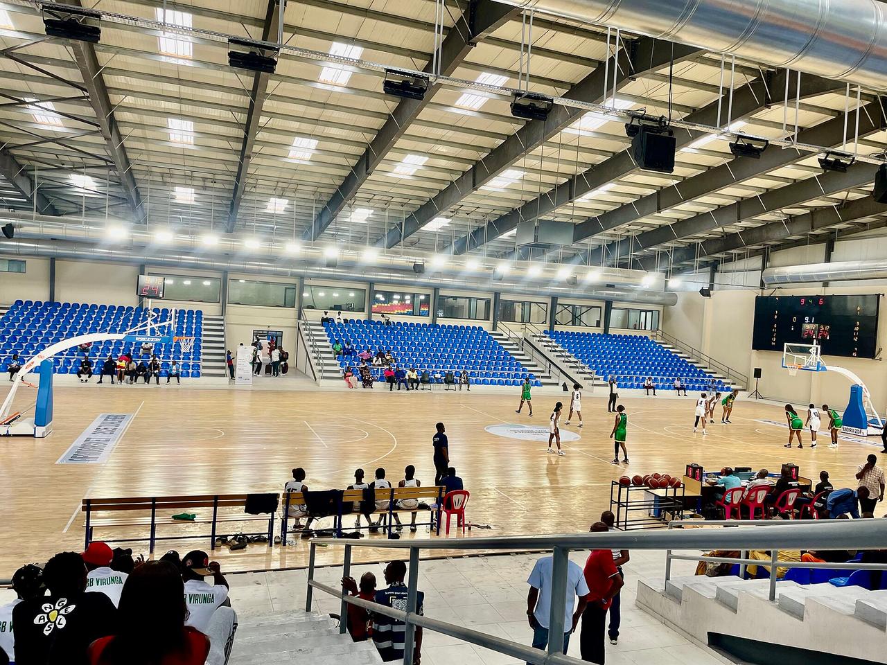 Vue intérieure du gymnase jumelé du Stade des Martyrs à Kinshasa : parquet de jeu clair, tribunes de sièges bleus partiellement occupées, tableau d'affichage électronique et charpente métallique de la halle, pendant une rencontre.