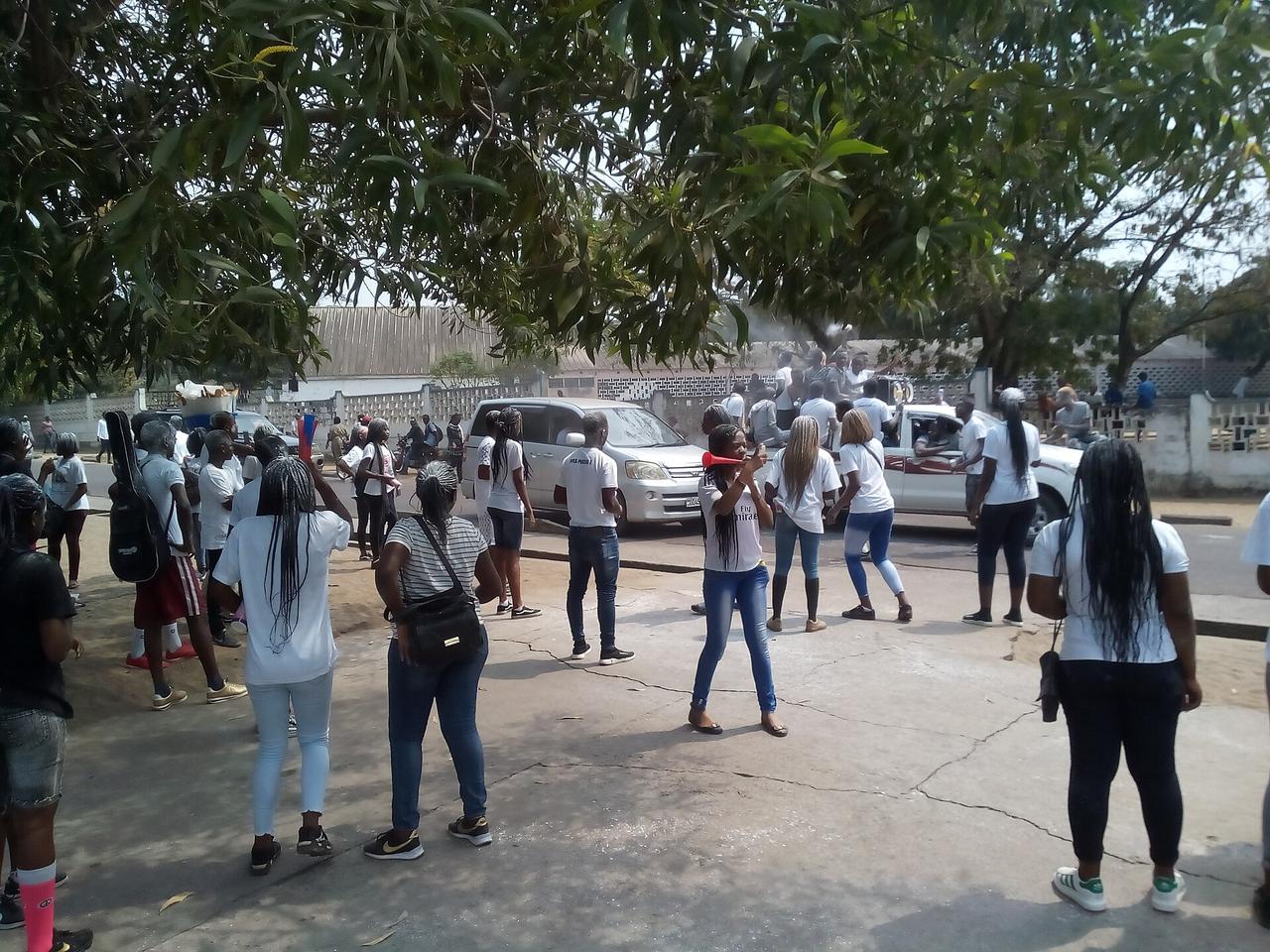 Des jeunes gens en t-shirts blancs rassemblés dans une rue de Kinshasa ; une jeune femme souffle dans une vuvuzela, d'autres sont montés à l'arrière d'une camionnette, sous des manguiers.