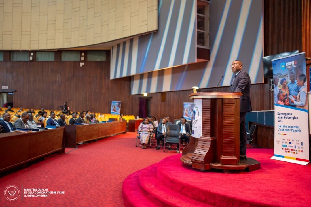 Guylain Nyembo s'exprime depuis un pupitre dans l'hémicycle du Palais du Peuple à Kinshasa, devant une assistance assise, à côté d'un kakémono du RGPH-2 portant le slogan « Se faire recenser, c'est bâtir l'avenir »
