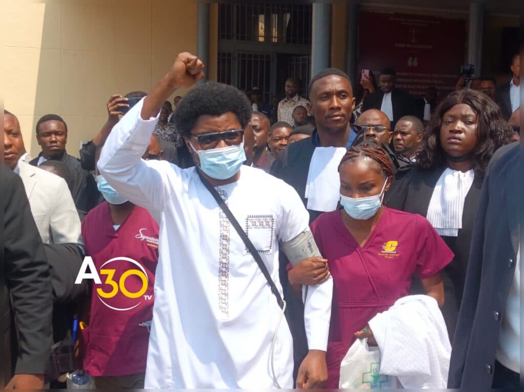 Constant Mutamba, le poing levé, portant un masque chirurgical et un brassard de tensiomètre au bras gauche, soutenu par une assistante médicale en tenue bordeaux, à la sortie de la Cour de cassation à Kinshasa, entouré d'une foule d'avocats en robe et de journalistes.