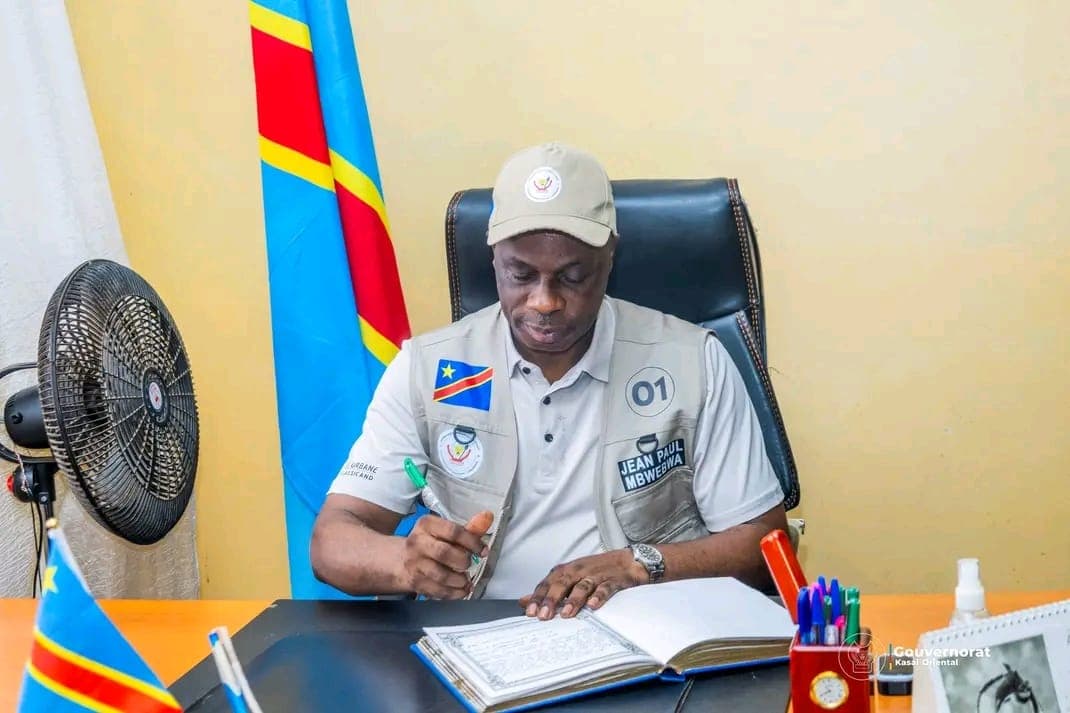 Le gouverneur du Kasaï-Oriental Jean-Paul Mbwebwa, casquette et gilet gris floqué de son nom et du numéro 01, signe un document à son bureau devant le drapeau de la RDC.