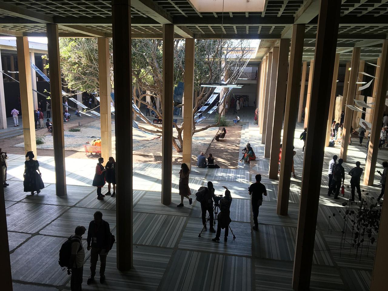 Vue plongeante sur la cour intérieure du site principal de la Biennale de Dakar : un vaste bâtiment ouvert à hautes colonnes, où des visiteurs circulent entre les installations, sous des arbres et des toiles suspendues.