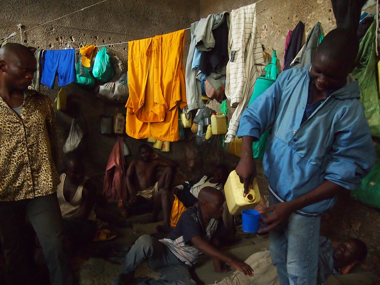 Intérieur d'une cellule surpeuplée de la prison de Bunia : des détenus assis et allongés à même le sol en terre battue, du linge suspendu à une corde le long du mur en béton, des bidons jaunes et des bouteilles d'eau accrochés à la paroi. Un homme debout verse de l'eau d'un bidon dans une tasse bleue.