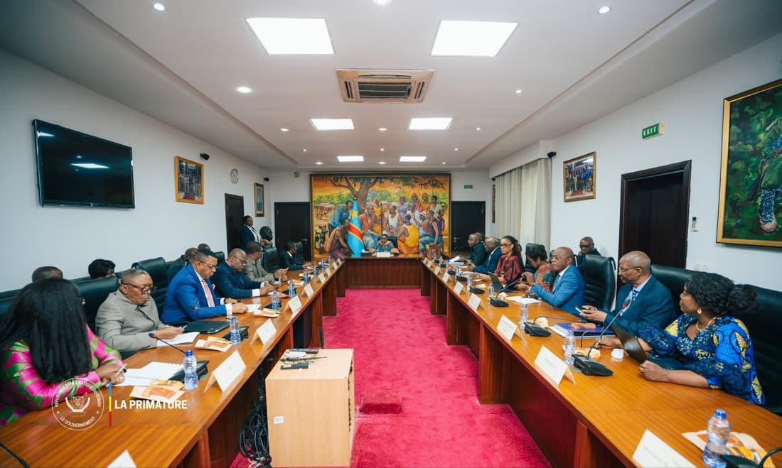 Vue d'une salle de réunion de la Primature à Kinshasa : membres du gouvernement, magistrats et responsables d'institutions assis de part et d'autre d'une longue table en U, face à la table de présidence placée devant une fresque murale aux couleurs du drapeau congolais.